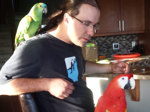 A man sits with a green parrot on his shoulder and a red parrot on his knee